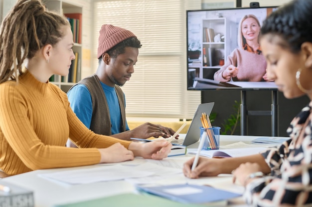A diverse group of students participating in an online class via a video conferencing platform. The students are engaged and interacting with the instructor on a split-screen. The setting is a mix of home offices and study spaces, showcasing the flexibility of online learning.
