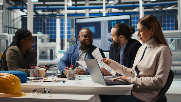 A diverse group of people, including engineers and business professionals, gathered around a table discussing solar panel designs, with laptops displaying energy efficiency data.